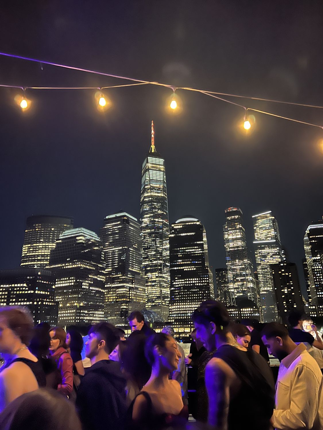 Crowd under string lights with the Manhattan skyline