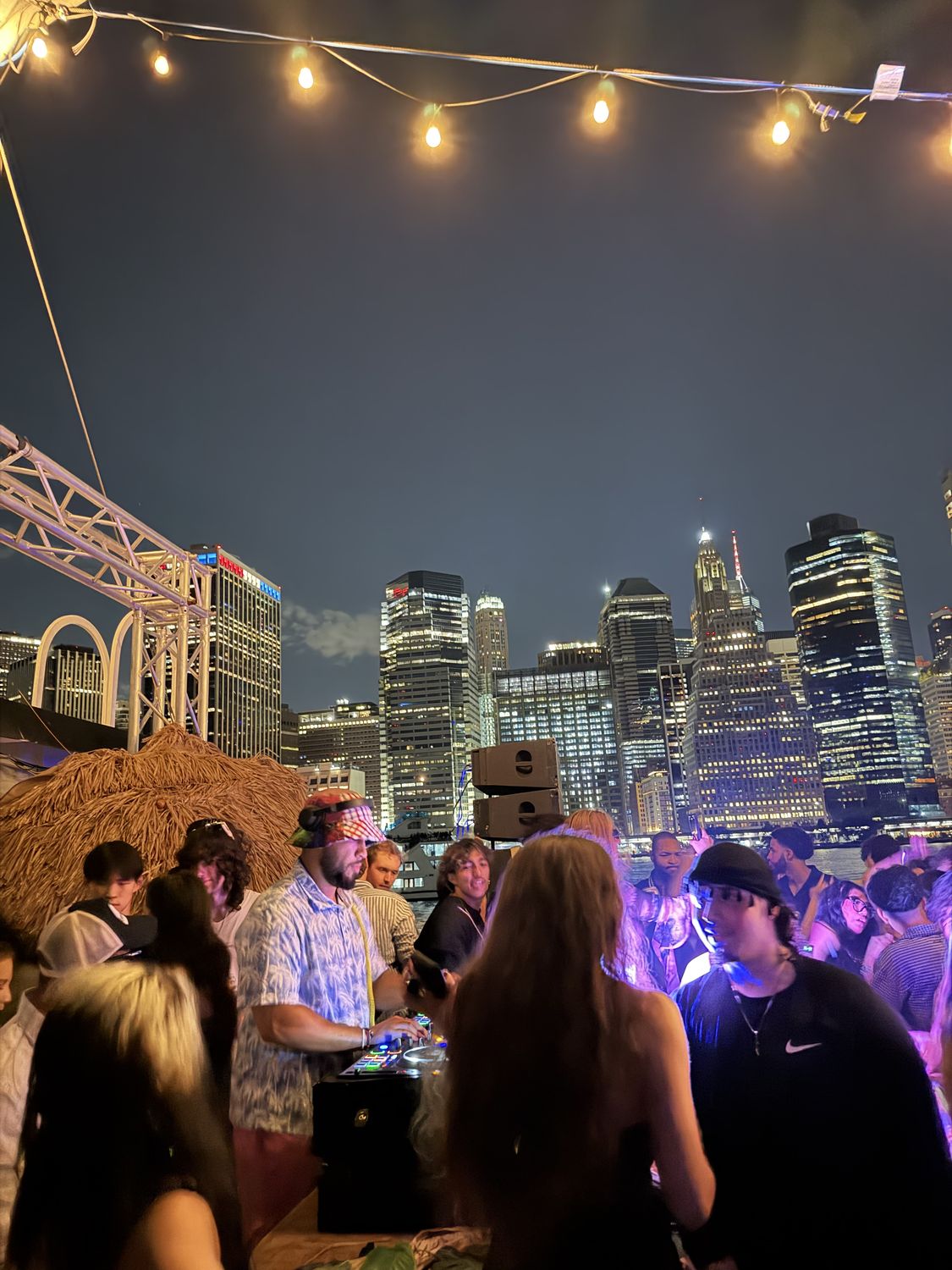 Crowd under string lights with the New York skyline at night